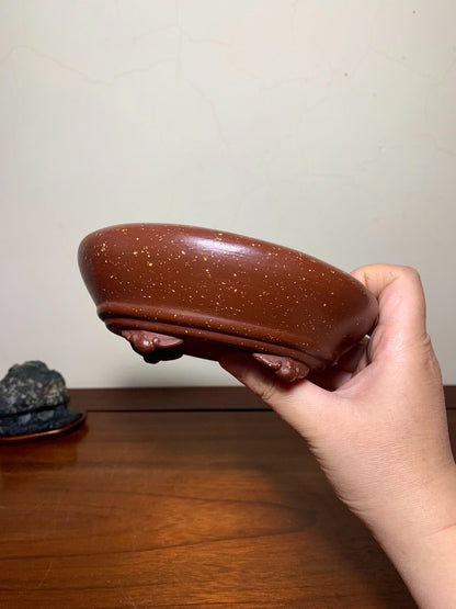 A hand holds a glossy brown ceramic pot with speckles and three small feet.