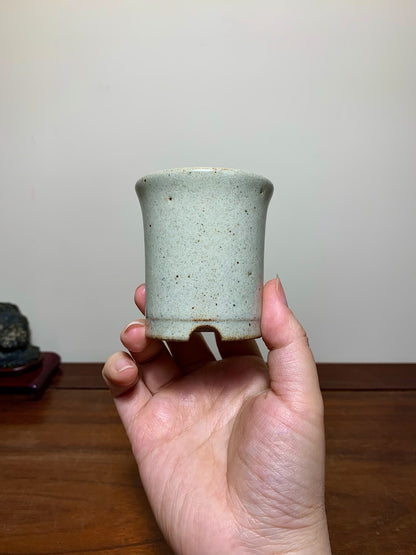 A hand holds a small, light green speckled glazed pot with a single drainage hole.