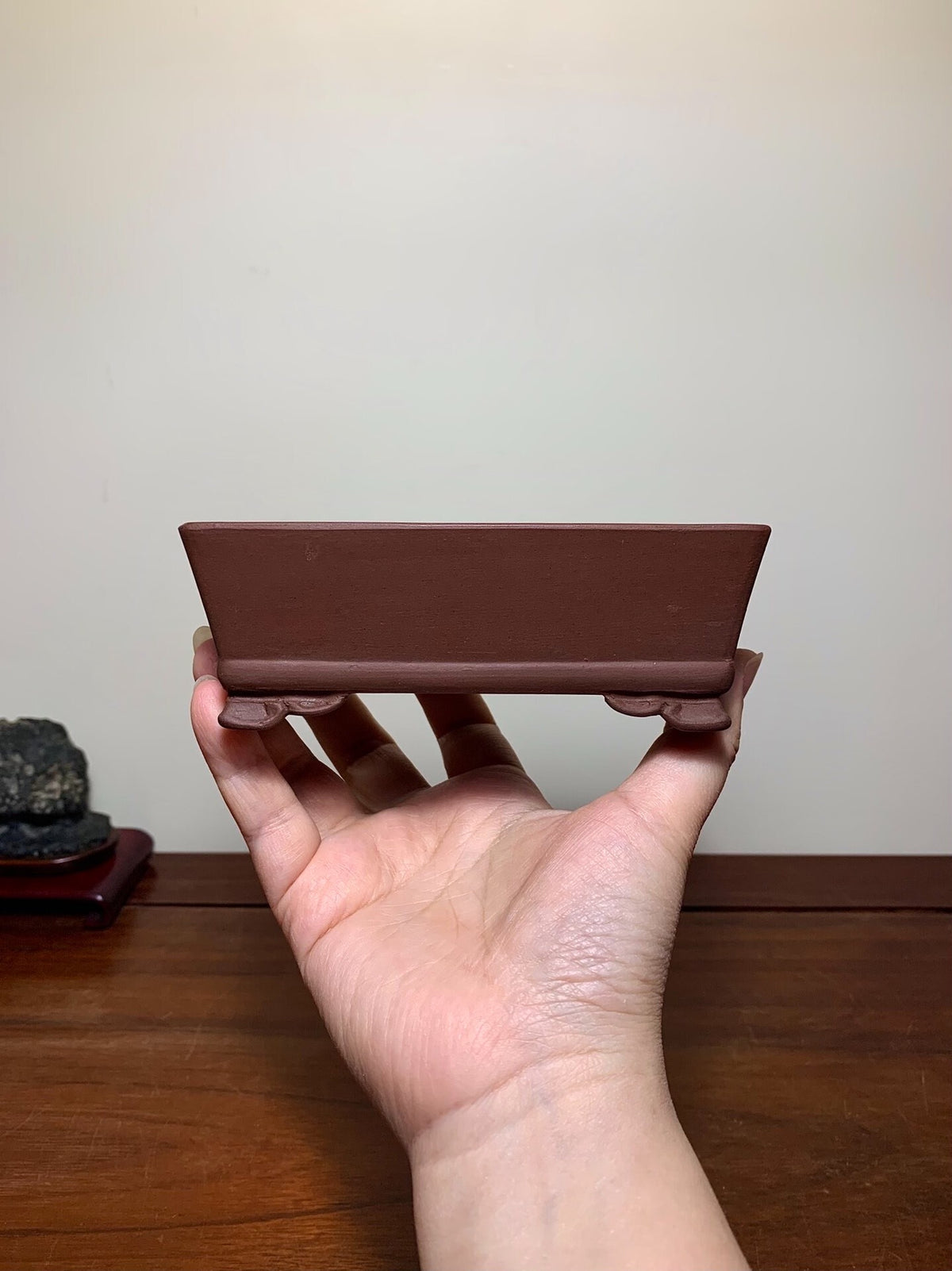 A hand holds a small, matte brown rectangular pot with two small feet, unglazed ceramic.