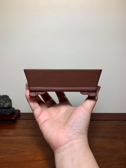 A hand holds a small, matte brown rectangular pot with two small feet, unglazed ceramic.