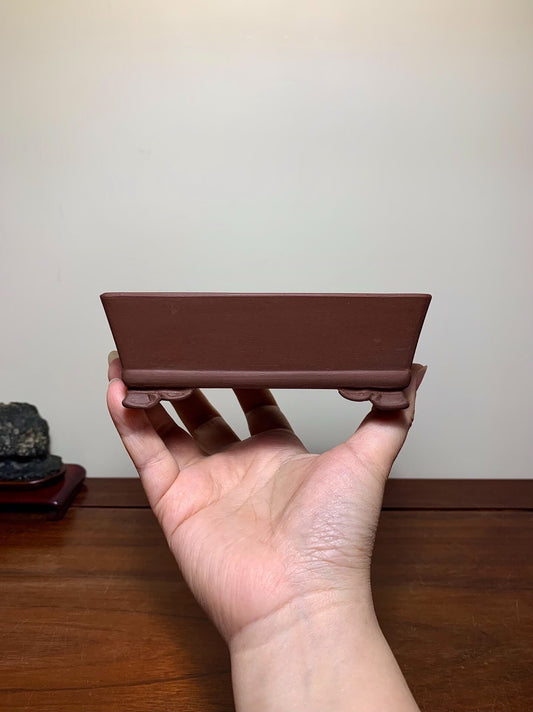 A hand holds a small, matte brown rectangular pot with two small feet, unglazed ceramic.