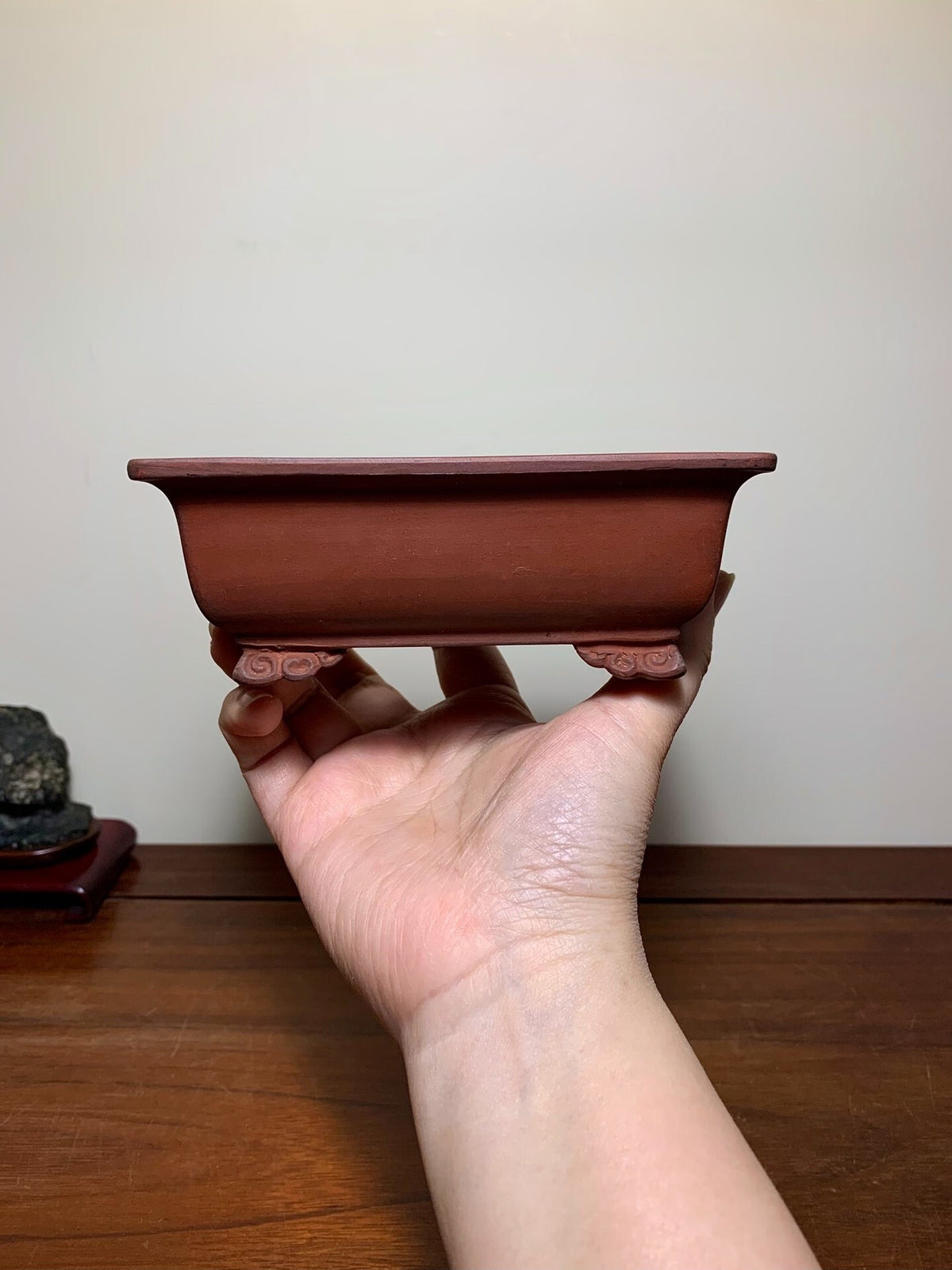 A hand holds a small, reddish-brown, rectangular ceramic pot with curved feet and a matte finish.