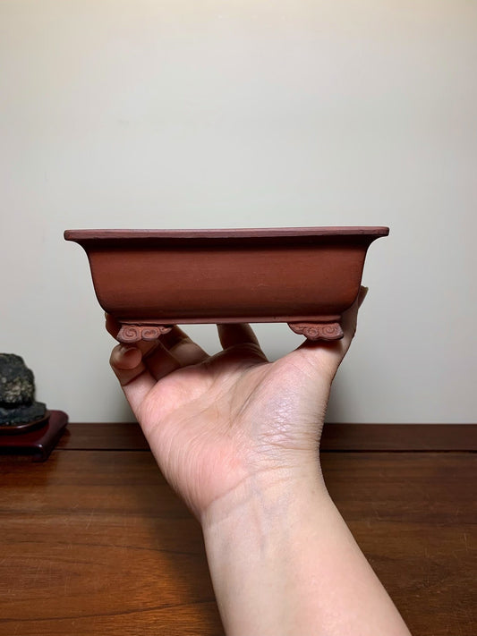 A hand holds a small, reddish-brown, rectangular ceramic pot with curved feet and a matte finish.