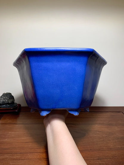 A hand holds a glossy, deep blue, hexagonal ceramic pot with flared sides and three feet.