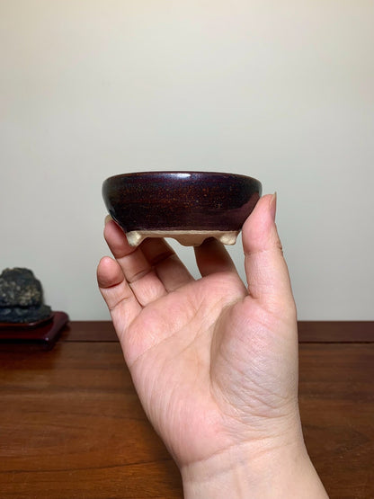 A hand holds a small, dark brown glazed pot with a speckled finish and three light-colored feet.