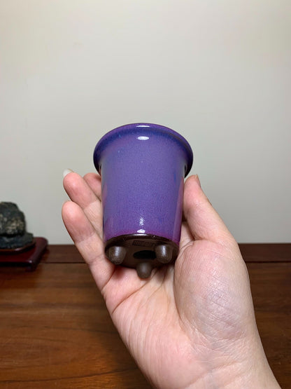 A hand holds a small, glossy purple ceramic pot with three dark brown feet.