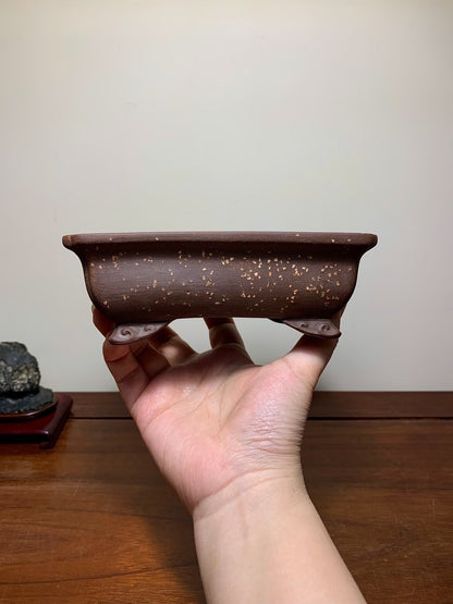 A hand holds a small, rectangular, brown unglazed ceramic pot with speckled surface and two small feet.