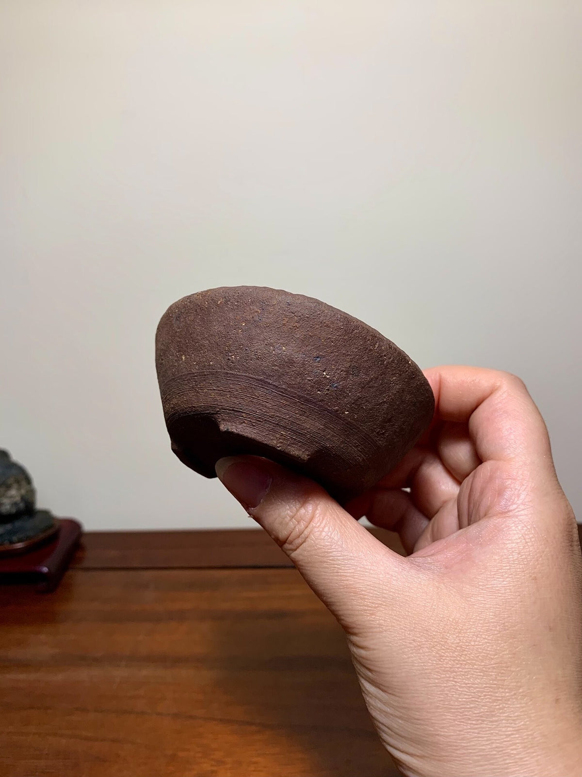 A hand holds a small, dark brown, unglazed ceramic pot with a rounded, shallow bowl shape.