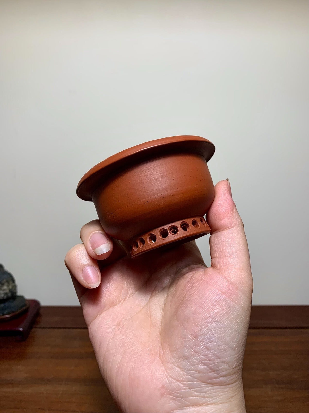A hand holds a small, unglazed terracotta pot with a flared rim and perforated base.