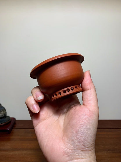 A hand holds a small, unglazed terracotta pot with a flared rim and perforated base.