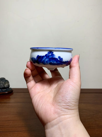 A hand holds a small white ceramic pot with a blue landscape painting and a blue rim.