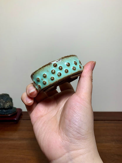 A hand holds a small, light blue glazed pot with brown rim and raised dots.