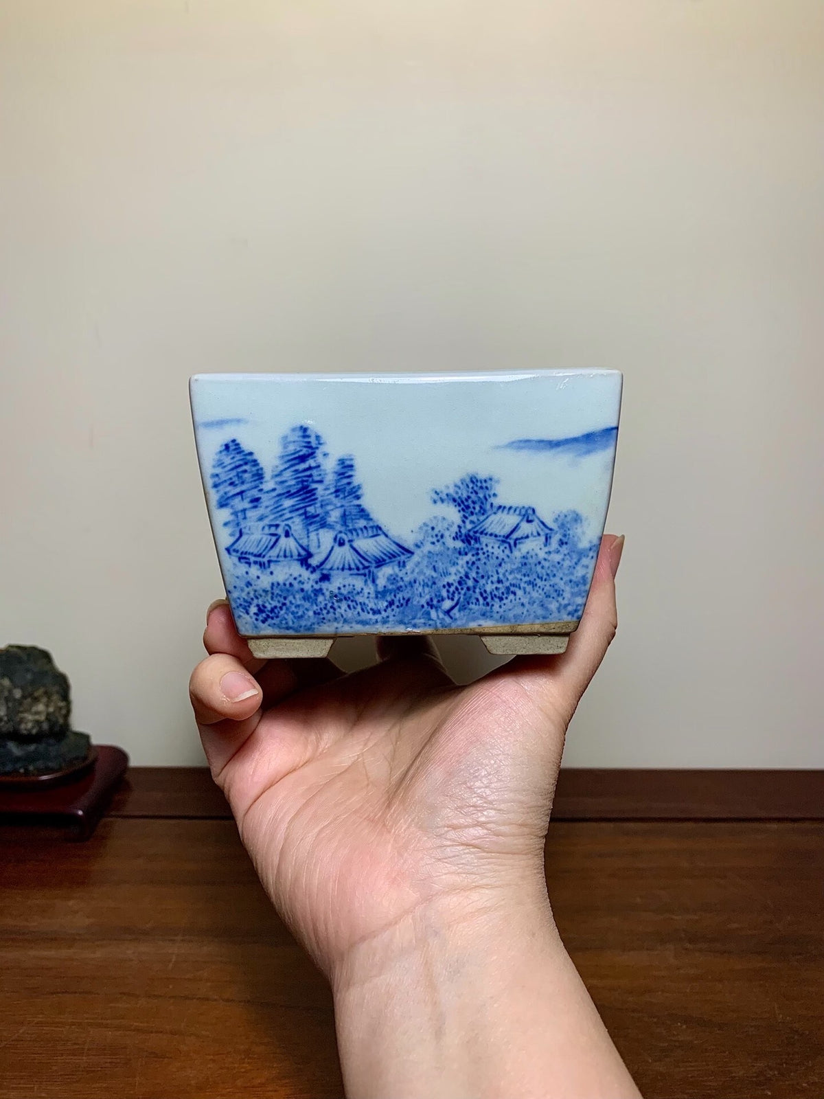 A hand holds a small, square blue and white ceramic pot with a landscape painting.
