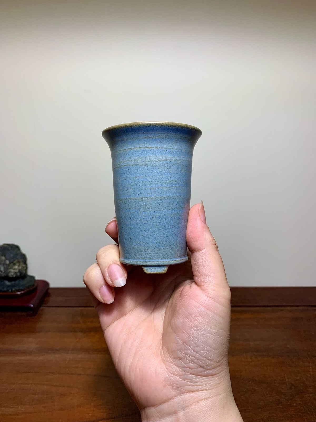 A hand holds a small, blue-glazed ceramic pot with a flared rim and subtle horizontal ridges.