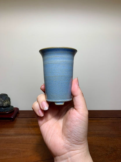 A hand holds a small, blue-glazed ceramic pot with a flared rim and subtle horizontal ridges.