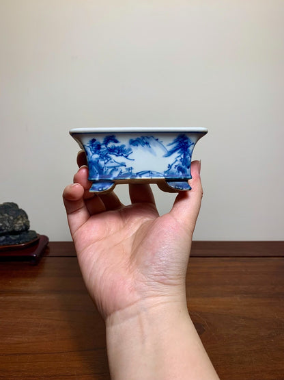A hand holds a small, rectangular blue and white glazed pot with painted landscape scenes.