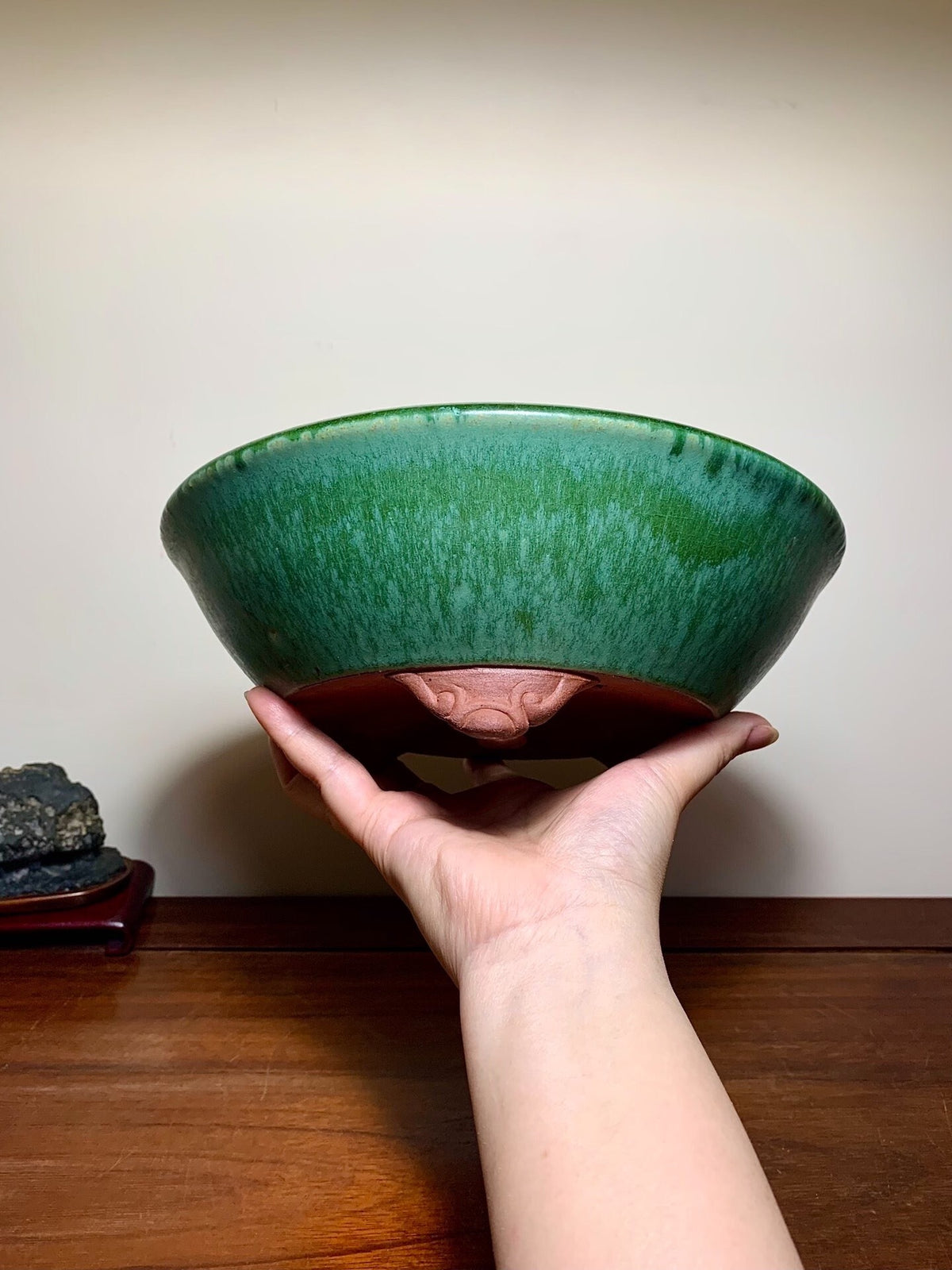 A hand holds a glossy green glazed pot with a brown rim and carved face detail.