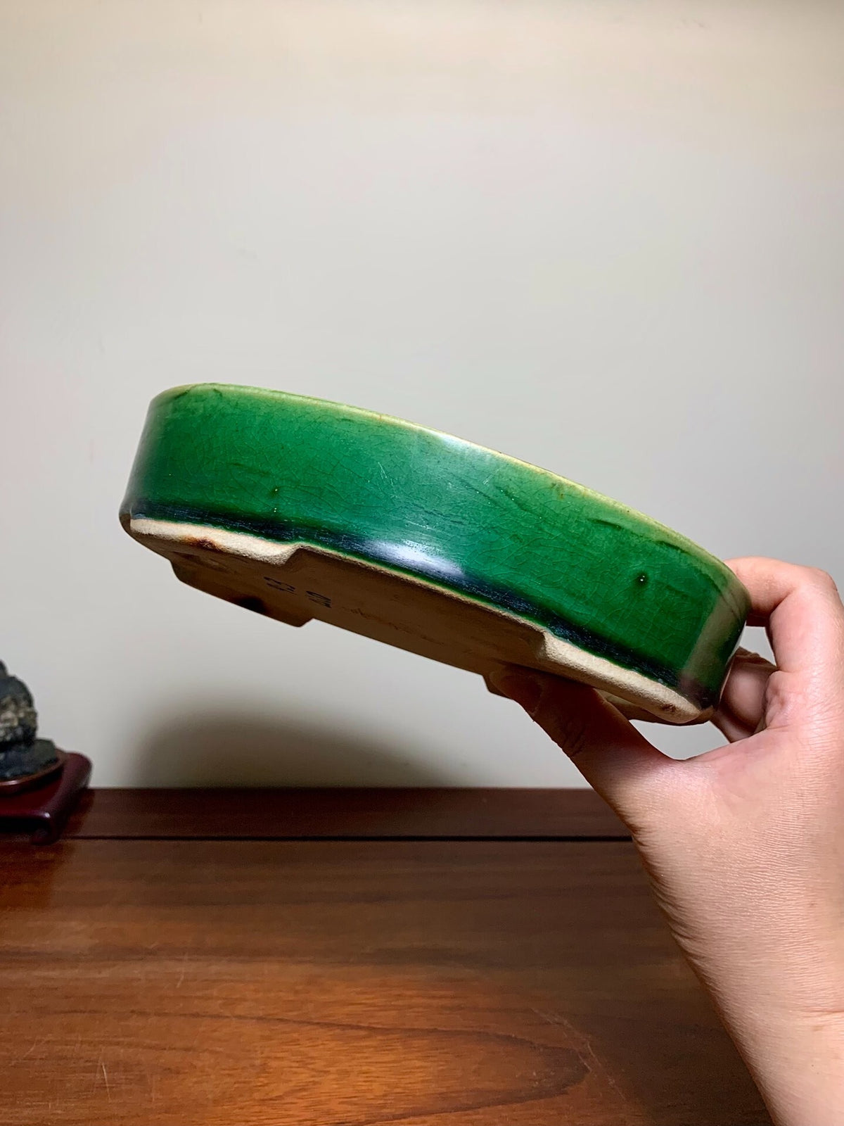 A hand holds a glossy green glazed ceramic bonsai pot with a wide rim and unglazed base.