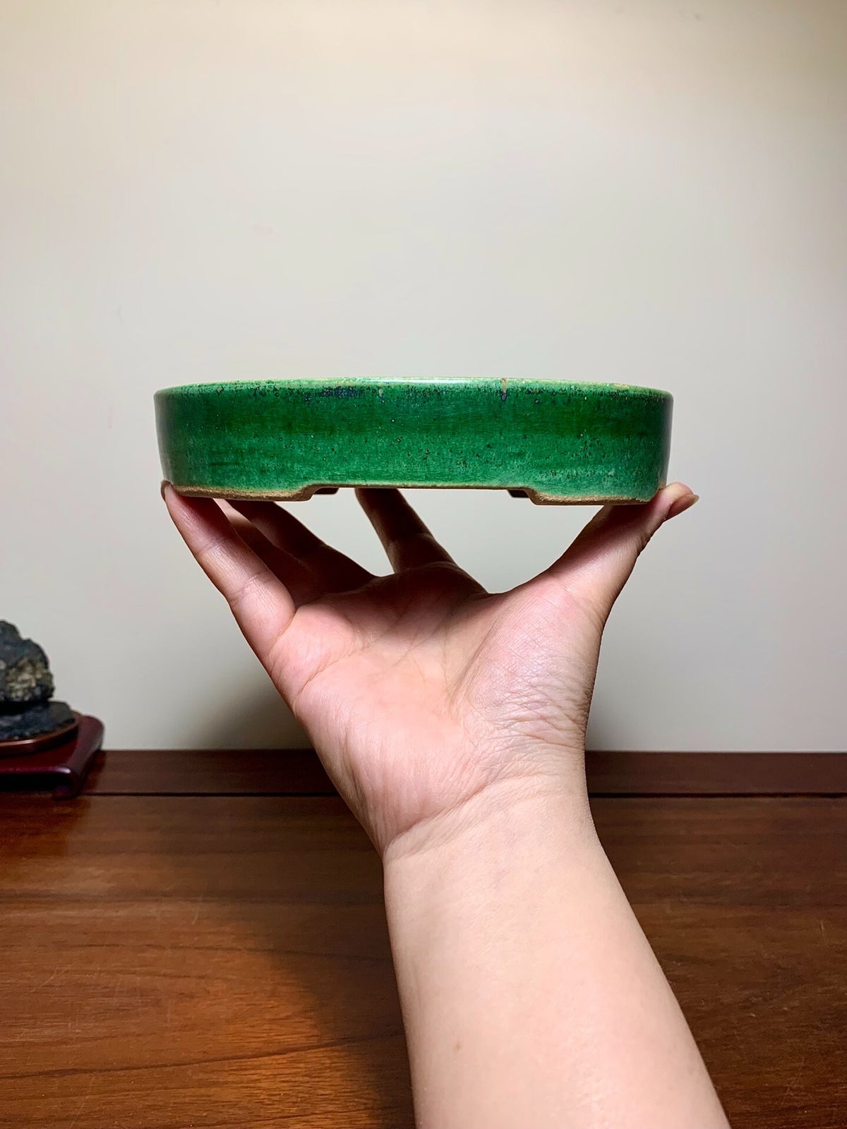 A hand holds a small, round, glossy green glazed pot with a slightly uneven rim.