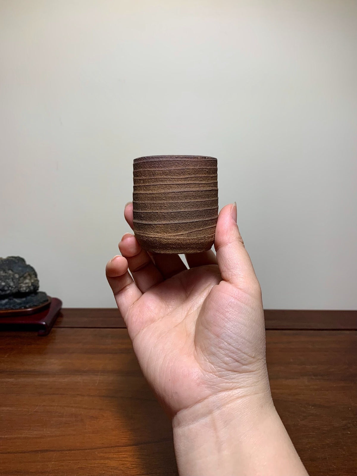 A hand holds a small, dark brown, matte-finished ceramic pot with horizontal ridges.