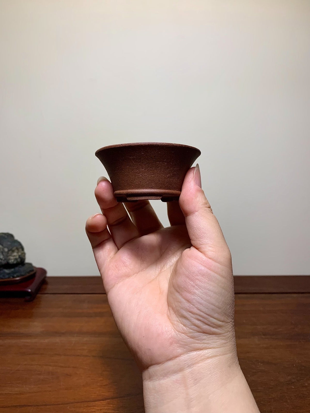 A hand holds a small, dark brown, unglazed ceramic pot with a flared rim and flat base.