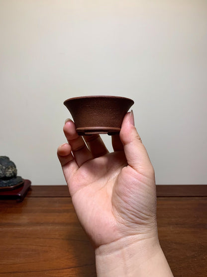 A hand holds a small, dark brown, unglazed ceramic pot with a flared rim and flat base.