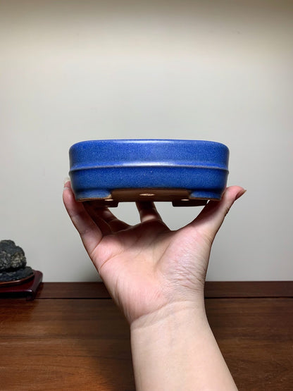 A hand holds a small, deep blue glazed rectangular pot with a matte finish and three wooden feet.