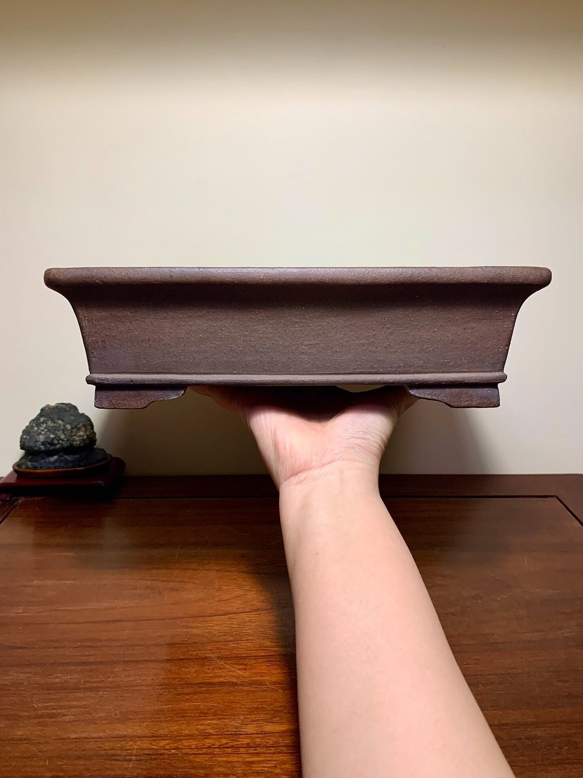 A hand holds a dark brown, rectangular, unglazed ceramic bonsai pot with a slightly curved rim.