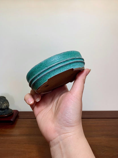 A hand holds a small, teal-glazed ceramic pot with three ridged rings around its rim.