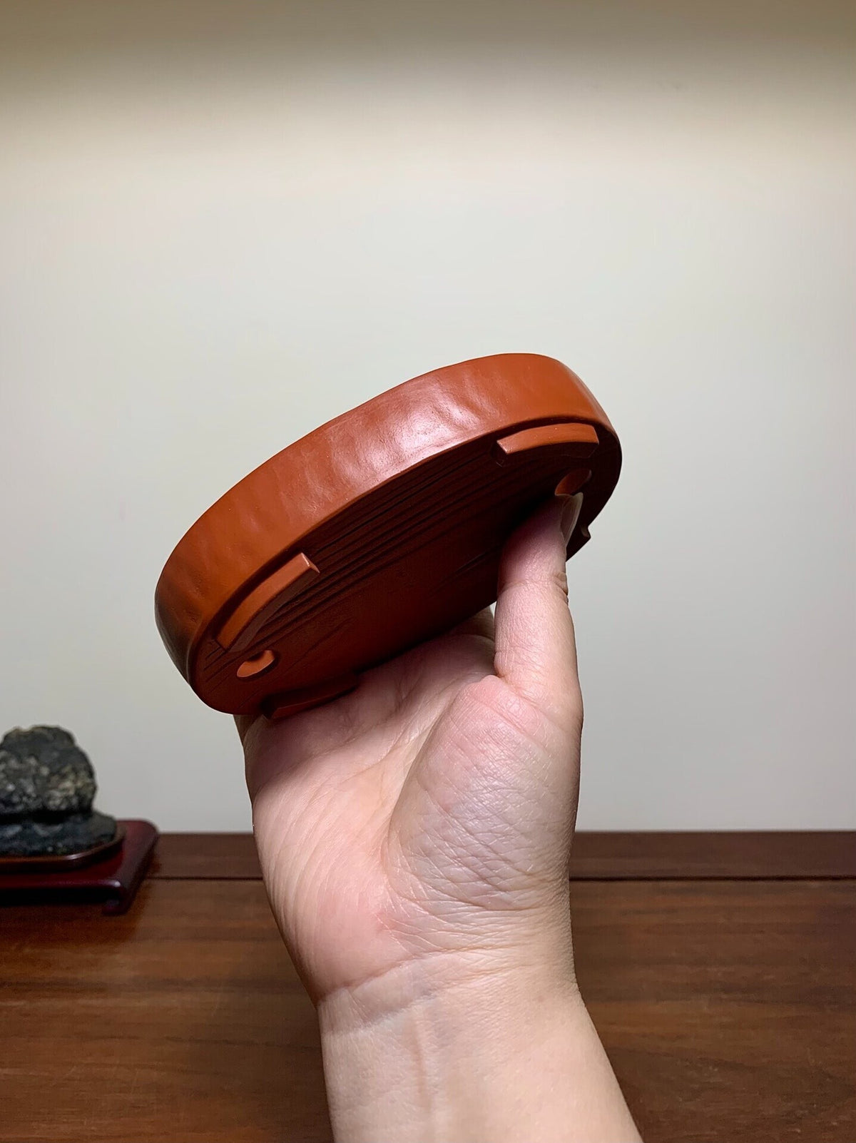 A hand holds a glossy, reddish-brown ceramic pot base with four small feet and concentric ridges.