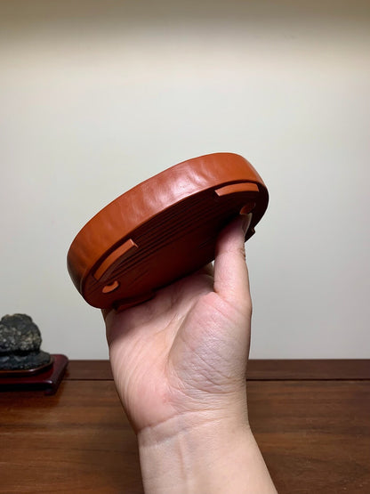 A hand holds a glossy, reddish-brown ceramic pot base with four small feet and concentric ridges.