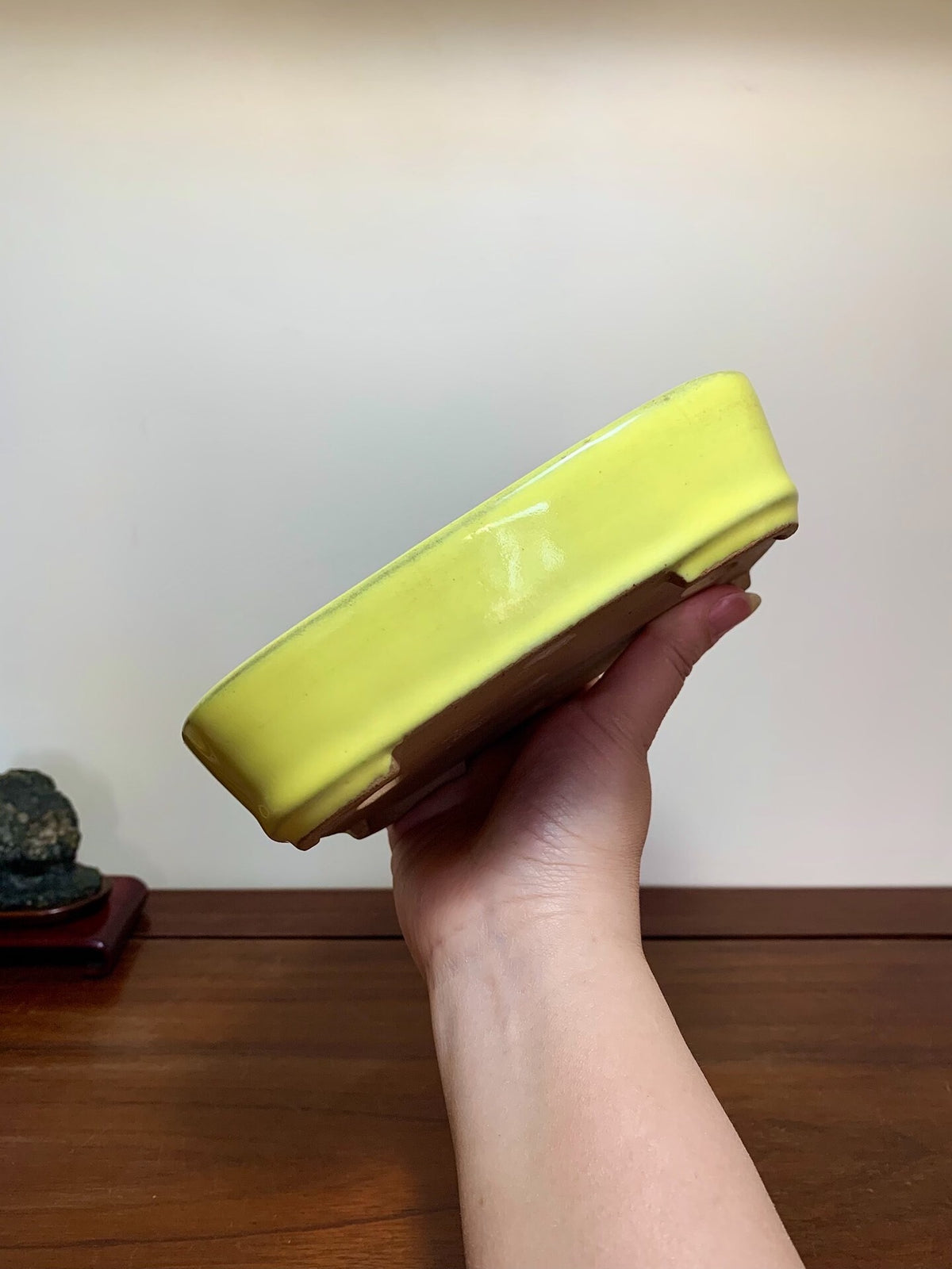A hand holds a glossy, pale yellow rectangular ceramic pot with a brown base.