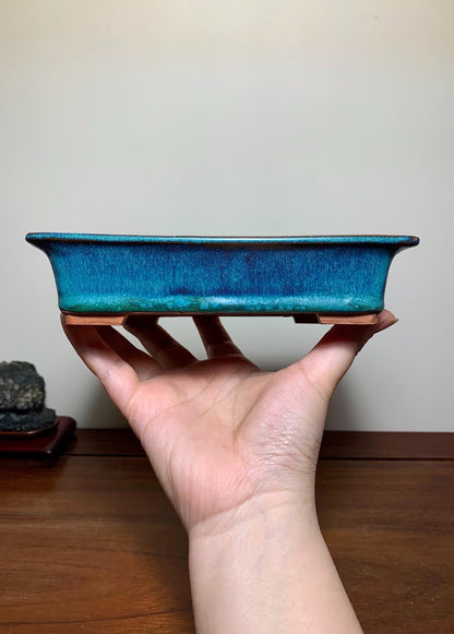 A hand holds a rectangular, glossy blue glazed pot with a textured surface and unglazed brown rim.