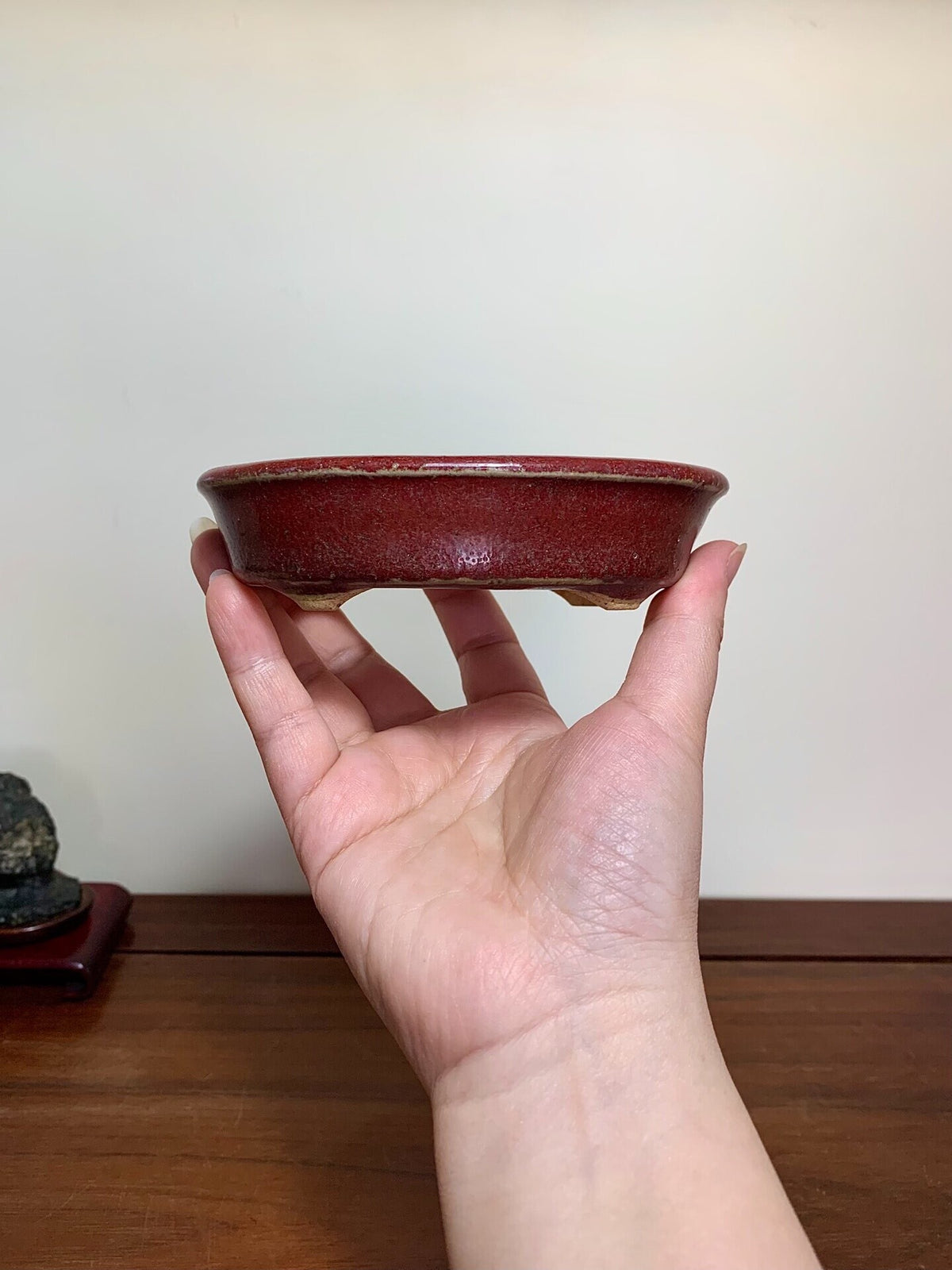 A hand holds a small, deep red glazed ceramic pot with a slightly uneven rim.