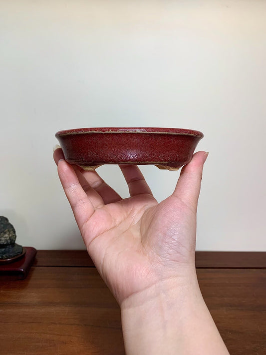 A hand holds a small, deep red glazed ceramic pot with a slightly uneven rim.