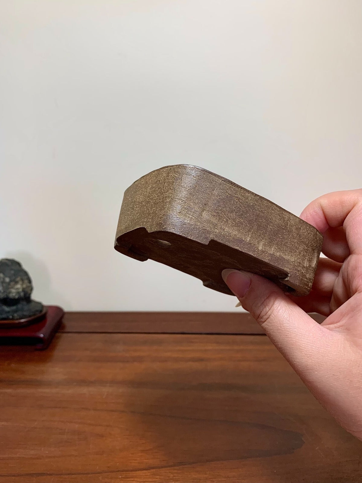 A hand holds a small, dark brown, matte-finished wooden pot with a curved, shallow bowl shape.