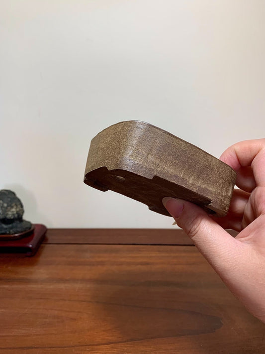A hand holds a small, dark brown, matte-finished wooden pot with a curved, shallow bowl shape.