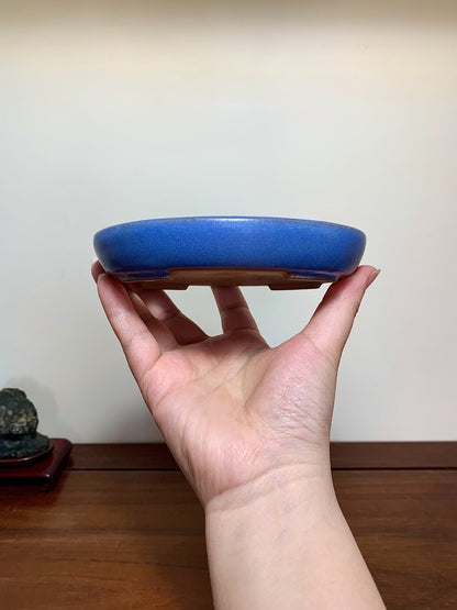 A hand holds a small, round, glossy blue glazed pot with a flat base.
