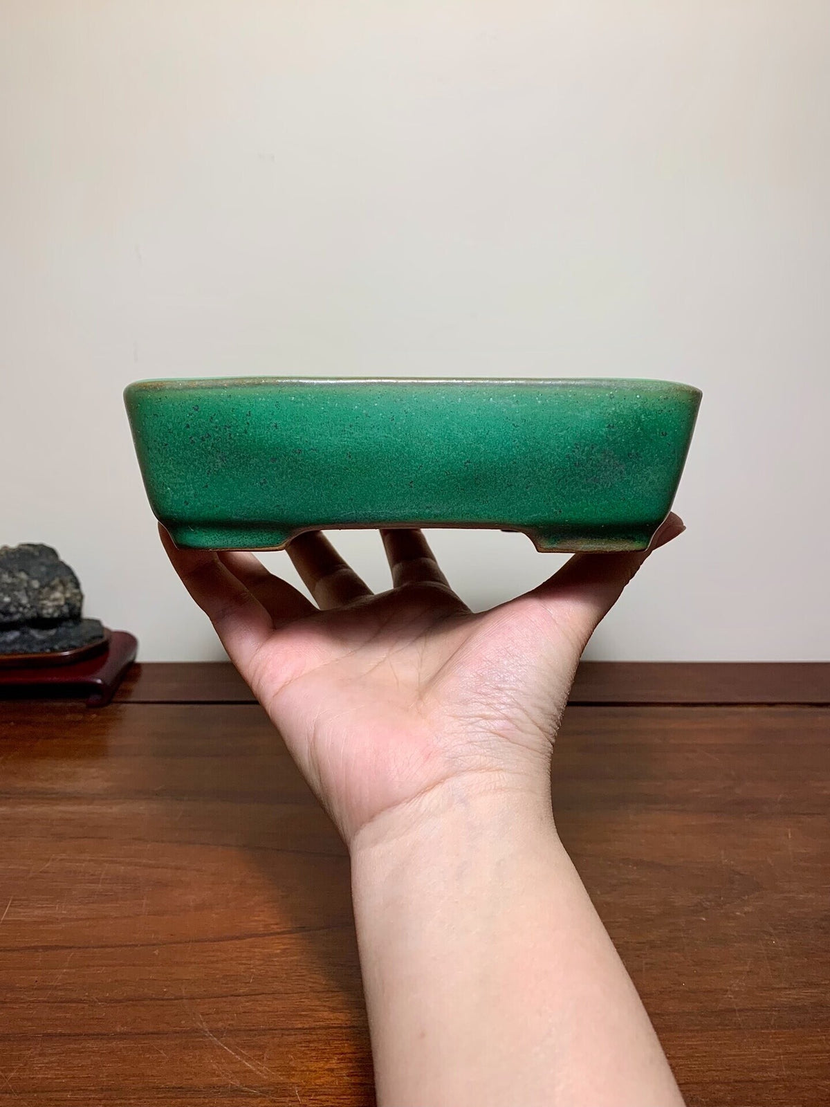 A hand holds a small, rectangular, glossy green glazed pot with a slightly tapered rim.