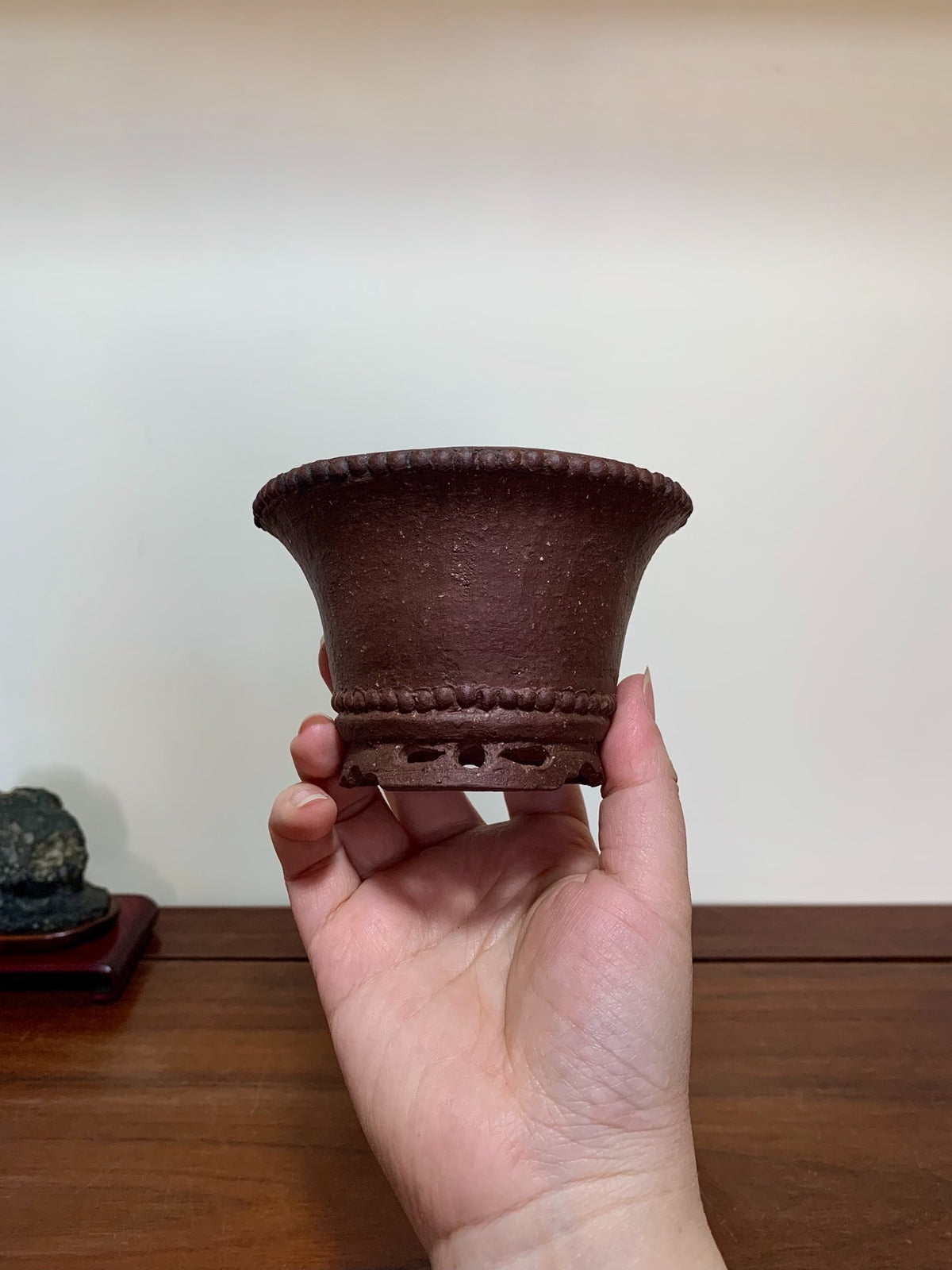 A hand holds a small, dark brown, unglazed ceramic pot with a flared rim and decorative bead trim.