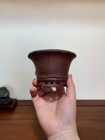 A hand holds a small, dark brown, unglazed ceramic pot with a flared rim and decorative bead trim.
