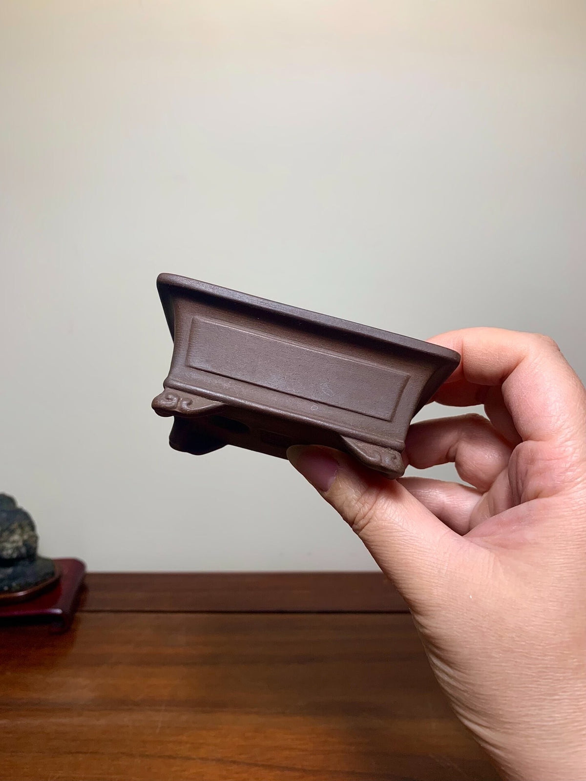 A hand holds a small, dark brown, rectangular ceramic pot with a matte finish and raised panel.