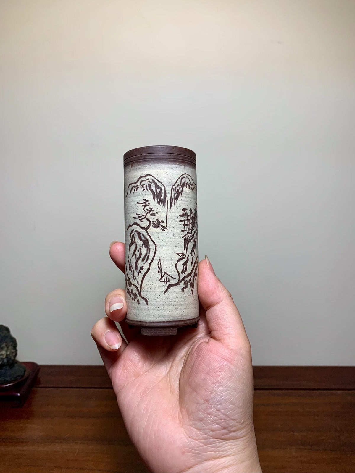 A hand holds a tall, cylindrical pot with a brown rim and greyish-white surface, featuring dark brown mountain and tree motifs.