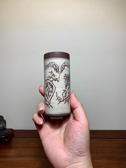 A hand holds a tall, cylindrical pot with a brown rim and greyish-white surface, featuring dark brown mountain and tree motifs.