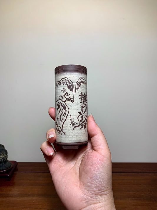 A hand holds a tall, cylindrical pot with a brown rim and greyish-white surface, featuring dark brown mountain and tree motifs.
