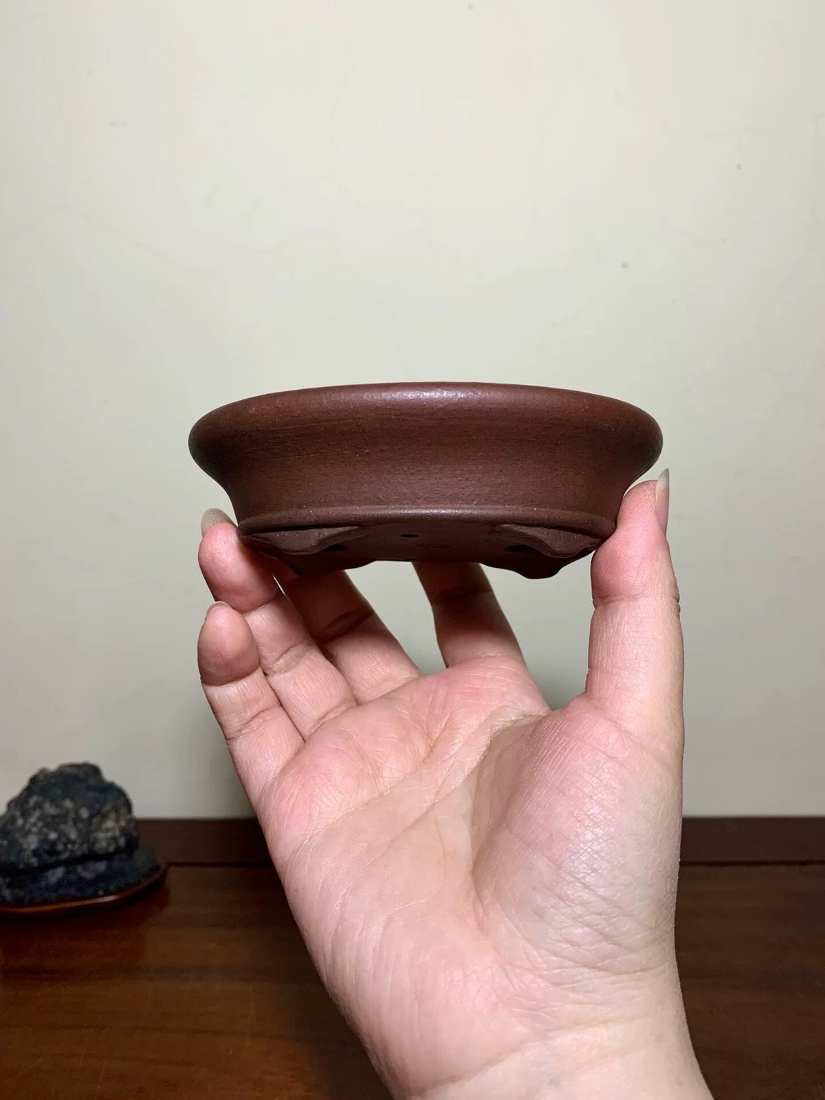 A hand holds a small, brown, unglazed ceramic pot with a wide rim and four feet.