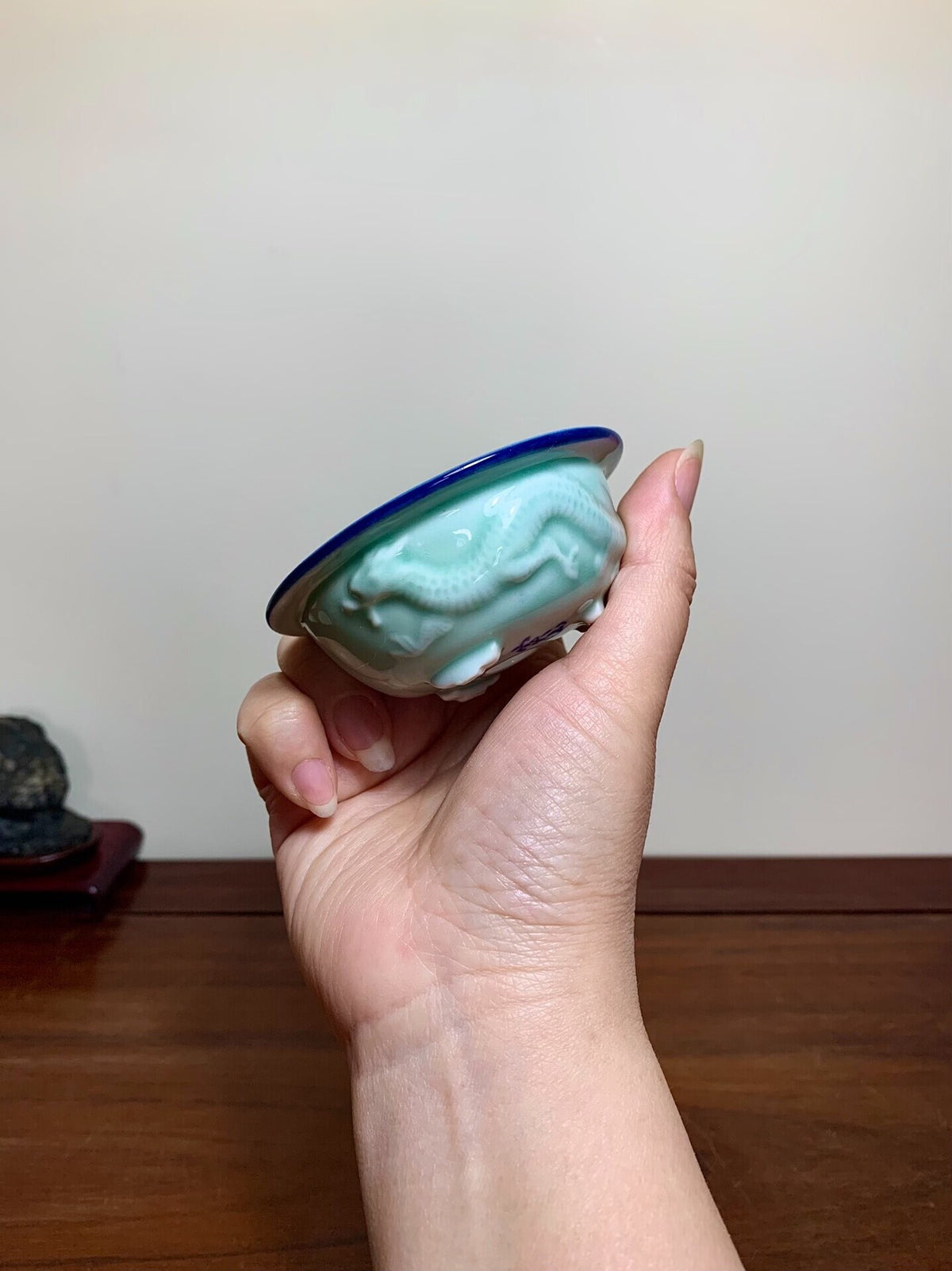 A hand holds a small, light green glazed pot with a blue rim and carved dragon design.