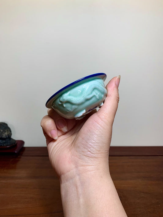 A hand holds a small, light green glazed pot with a blue rim and carved dragon design.
