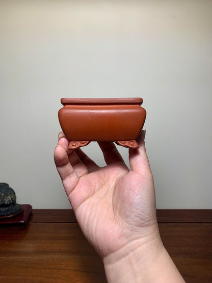 A hand holds a small, reddish-brown, rectangular glazed pot with four footed supports.