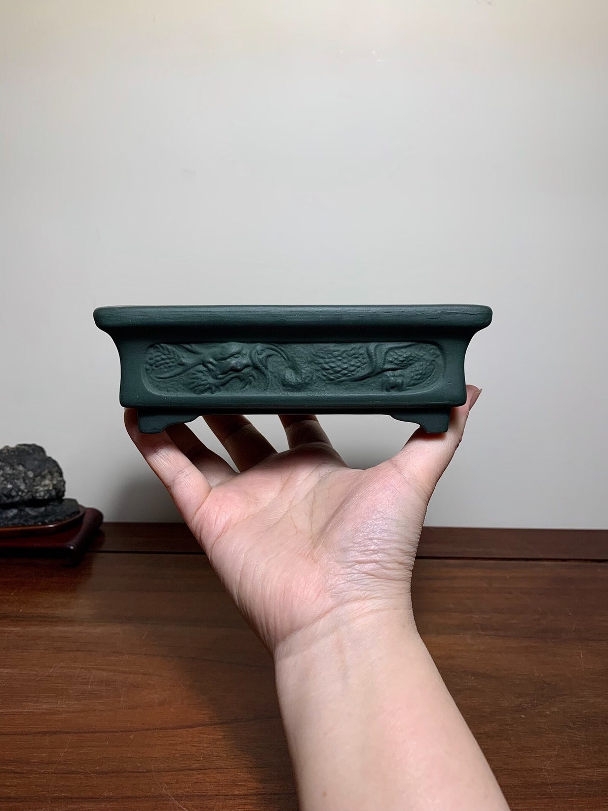 A hand holds a dark green, rectangular bonsai pot with a carved dragon design.